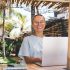 Positive young female freelancer in glasses and casual outfit working online on project using laptop sitting in gazebo near villa and palms